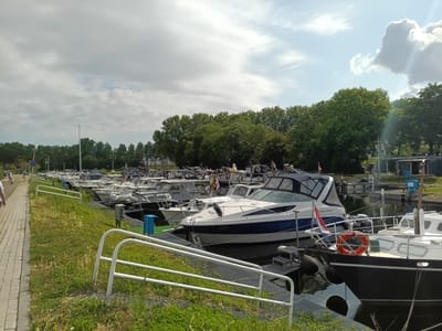 The image shows a marina in Zwartewaal, Netherlands, with numerous boats docked along a waterfront. A paved walkway runs along the left side of the frame, with a grassy embankment sloping down to the water. Several people are visible walking on the pathway, including one person on a scooter. A row of boats, varying in size and design, are moored in the water, with their masts and decks clearly visible. Some of the boats are sleek, modern vessels, while others appear to be more traditional motorboats. In the background, a dense line of trees forms a natural barrier, and beyond that, buildings and infrastructure related to the marina are discernible. The sky is partly cloudy, with patches of blue and white, suggesting fair weather. A sign with Dutch text, possibly indicating the location or a marina facility, is visible on one of the buildings in the background. The scene depicts a typical day at a busy harbor, with boats ready for use and people enjoying the waterfront.