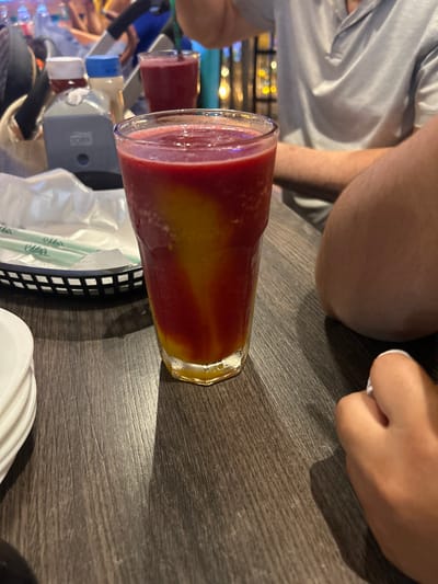 The image captures a close-up, angled view of a layered beverage in a tall glass, set on a dark wooden table. The drink exhibits vibrant colors, with a deep red layer at the bottom and top, separated by a bright yellow or orange layer in the middle, creating a striking visual effect. Condensation on the glass suggests the drink is cold.

Around the main subject, several elements contribute to the scene. To the left and slightly behind the prominent glass, another similar red beverage in a glass is partially visible, indicating multiple drinks are present. A small, black, rectangular basket sits on the table, containing neatly folded white napkins and several green plastic straws. The word "Eddy's" is clearly visible in white lettering, repeated along the length of the green straws. In the bottom right corner, a stack of white plates or bowls is partially seen.

Parts of at least two individuals are visible in the frame. On the upper left, a person wearing a light grey t-shirt is seen, with their arm resting on the table. Further to the top right, a hand with light skin and white painted fingernails is visible, also resting on the table. Another bare limb, likely an arm or leg, with light brown skin tone, is visible in the upper central portion of the image.

The setting appears to be an indoor commercial establishment, possibly a restaurant or cafe, given the type of table, drinks, and background elements. Blurry lights in the distant background suggest an ambient evening or nighttime atmosphere. The overall image is rotated approximately 90 degrees counter-clockwise.