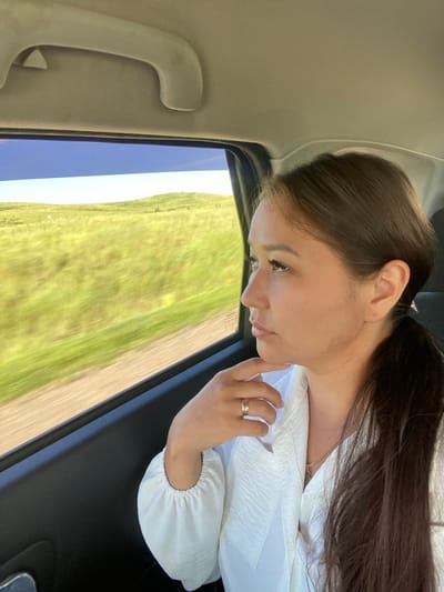 A young woman with dark hair pulled back in a ponytail sits in the back seat of a vehicle, looking out the window. She wears a white shirt with ruffles on the sleeves and a ring on her finger. The window reveals a blurred landscape of green hills and a blue sky, suggesting the vehicle is in motion. The interior of the car is visible, including a handle on the ceiling and the dark upholstery of the door. The scene appears to be in a rural area.