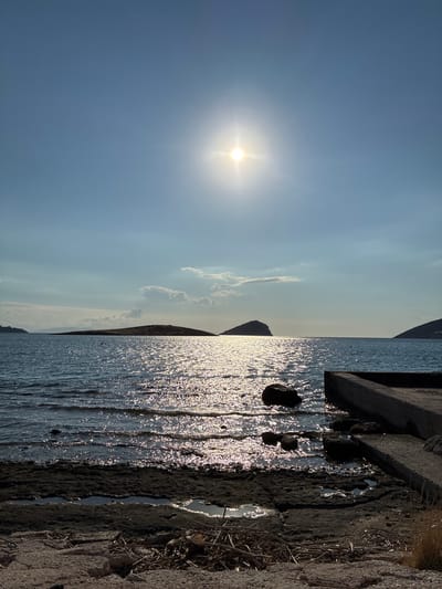 The image depicts a seaside scene in Limenas Markopoulou, Greece, on a clear, sunny day. The sun, a bright orb with visible rays, dominates the upper portion of the sky, casting a shimmering reflection across the rippling water. In the middle ground, several small, rocky islands are visible against the horizon. In the foreground, a concrete structure, likely a pier or a breakwater, extends into the water from the right side of the frame. The shoreline is composed of dark, uneven rock and sand, with some puddles of water and scattered dry vegetation. The overall atmosphere is serene and tranquil, characteristic of a coastal landscape.