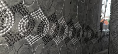 The image is a close-up, angled shot of a sheer black and white patterned curtain. The pattern consists of diamond shapes made of small stitched dots, alternating between black and white threads. Interspersed throughout the curtain are swirling, decorative white and black embroidered lines and small floral-like embellishments. The fabric is translucent, with a shimmering, metallic-like background pattern visible underneath the embroidery. To the right, a window is partially visible, showing a blurred outdoor scene with buildings and greenery, suggesting an urban or suburban environment. The curtain appears to be indoors, possibly in a residential setting. The lighting suggests it is daytime. There are no people or specific actions depicted.