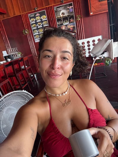 A selfie captures a young woman with curly dark hair and light skin, wearing a red halter top and a pearl and gold necklace that spells "Anita". She is holding a gray cup in her right hand and has several bracelets on her wrist. The background suggests a restaurant or bar setting in Preah Sihanouk, Cambodia, with wooden walls, red chairs, and a menu poster visible on the wall. A white fan is positioned to her left, and a tropical plant is seen in the background.