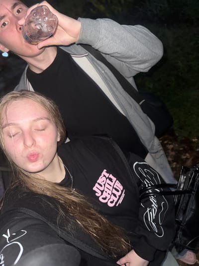 In this low-light, outdoor image, two individuals are partially visible. In the foreground, a young woman with blonde hair pulled back is facing the camera with her eyes closed and lips pursed in a kissing gesture. She is wearing a black long-sleeved shirt with pink text that reads "JUST CHILL CLUB." A silver necklace with a cross pendant is visible around her neck. Behind her, a person, likely male, is seen from the chest up, drinking from a clear plastic water bottle. They are wearing a grey hooded jacket over a black t-shirt. The background is dark, suggesting nighttime, with some blurred greenery and fallen leaves on the ground, indicative of a park or wooded area. The overall scene appears casual and informal. The location in Ryabinino, Russia, is not visually evident from the image itself.