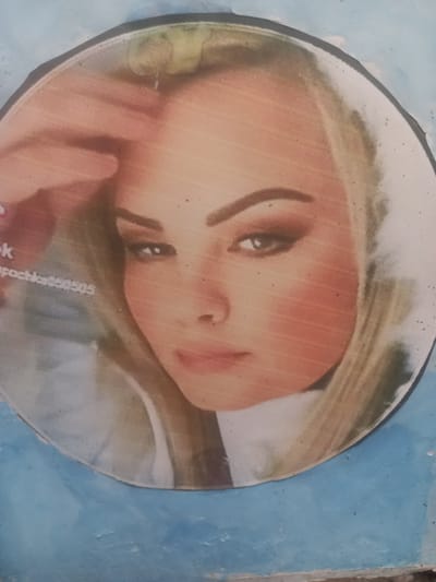 The image shows a close-up of a circular photograph of a woman's face. The photograph is mounted on a rough, light blue surface that appears to be a wall. The woman has blonde hair, and her face is turned slightly to the side. She is wearing makeup, including eyeliner and eyeshadow. In the lower left corner of the photograph, there is some text that is partially obscured. The photograph is slightly faded and has some speckling. The overall impression is of a worn, personal item displayed in an outdoor or semi-outdoor urban environment. There are no visible landmarks that would specifically place this image in Maloye Lugovoye, Russia, other than what is provided in the prompt.