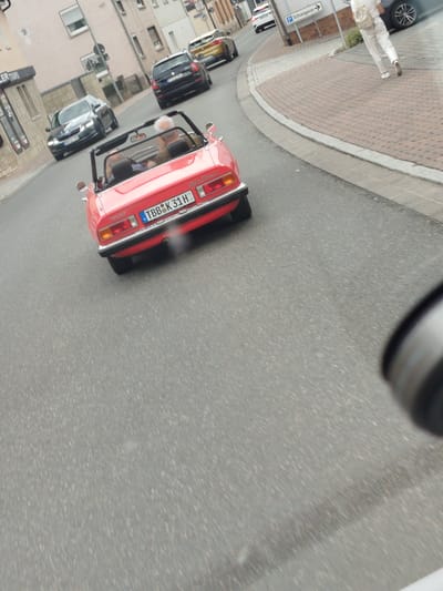 A red convertible classic car, possibly an Alfa Romeo Spider, is driving on a street. The car's license plate reads "TBB K 31 H". A man with white hair is in the driver's seat. In the background, other cars are visible, including a black Skoda and a yellow Audi. A person in white clothing is walking on the sidewalk. The scene appears to be in a town or city, with buildings lining the street. There is a signpost with German text that includes "Ochsensgasse". The weather appears to be overcast. The time of day is indeterminate but likely daytime.