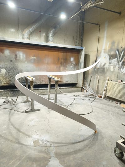 This image captures the interior of an industrial workshop or finishing area, located in Negorci, North Macedonia. The primary subject is a long, light-colored, thin strip of material, possibly wood or composite, bent into a horizontal serpentine curve. This strip is supported at two points by identical grey metal stands, each featuring a narrow wooden top. The far right end of the curved strip rests on a small, orange-brown wooden block directly on the concrete floor.

In the background, a prominent industrial spray booth or ventilation system dominates the rear wall, characterized by a dark blue upper section and a rust-colored lower panel, heavily stained with overspray. Above the booth, two silver, corrugated flexible ventilation ducts are visible, curving upwards towards the ceiling where multiple bright overhead lights are mounted. The walls exhibit significant staining and paint splatters in various colors, indicative of a working environment.

The concrete floor is soiled and worn, showing numerous paint splatters, scuff marks, and areas of discoloration. Several black electrical cables and air hoses are coiled and spread across the floor. To the right, a white panel radiator is affixed to the wall, with various pieces of wood and debris leaning nearby. A dark metal wheeled cart is partially visible in the lower right corner. No individuals are present within the frame.
