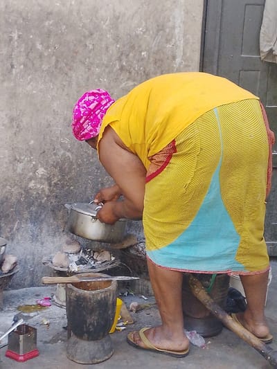 A woman wearing a yellow top and a yellow and blue patterned wrap bends over a cooking area outside. She has a pink headscarf and is holding a pot over a fire. The cooking area includes a mortar and pestle and various other pots and utensils. The wall behind her is stained and discolored. She is wearing sandals.