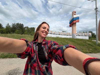 In Nikolaevka, Russia, a woman takes a selfie in front of a welcome sign. The sign, featuring the Russian flag colors and coat of arms, reads "VLADIVOSTOK." The woman is wearing a red and blue plaid shirt over a black top. The environment is an outdoor setting with a grassy hillside, trees, and a cloudy sky. A lamppost and a road are also visible.