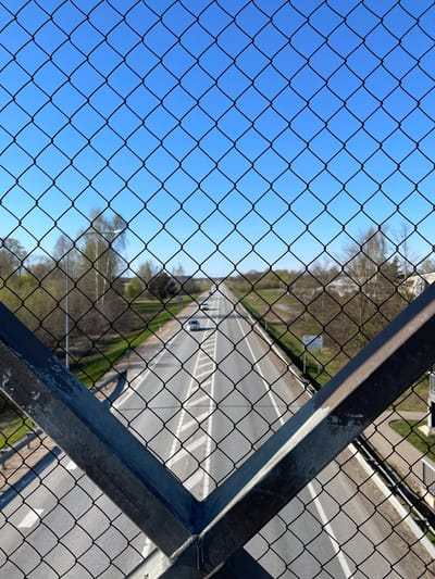 The image depicts a highway view through a wire fence and metal structure, possibly from a pedestrian bridge or elevated platform. The scene includes a section of highway with multiple lanes, white lane markings, and vehicles traveling on the road. The surrounding environment appears to be a wooded area with trees visible along the sides of the road. The sky is bright blue. The location is in Ādaži, Latvia.