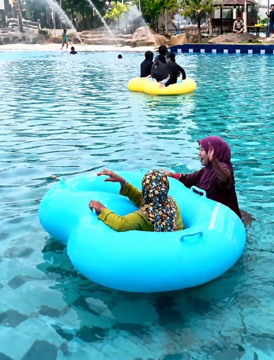 The image depicts a scene at a water park, likely in Eampat Kwalamencirim, Indonesia. In the foreground, two individuals are seated in a large, bright blue inflatable tube. One person, facing away from the camera, wears a patterned headscarf and a green long-sleeved shirt. Their hands are visible resting on the edge of the tube. To their right, another person wearing a purple headscarf and a dark top is partially visible, facing left and appearing to wave or reach out.
In the mid-ground, a group of at least three individuals, all wearing dark clothing, are seated in a yellow inflatable ring. They appear to be floating in the water.
The background features a water attraction with several water jets spraying water into the air, creating a misty effect. Several palm trees and other foliage are visible, along with rock formations. In the far background, a rustic wooden structure with a roof, possibly a seating area or shelter, is present. Other figures, including children and adults, are scattered throughout the water and on the paved areas surrounding the pool. The water is a clear, bright blue, with visible ripples and reflections. The overall atmosphere suggests a recreational setting on a sunny day.