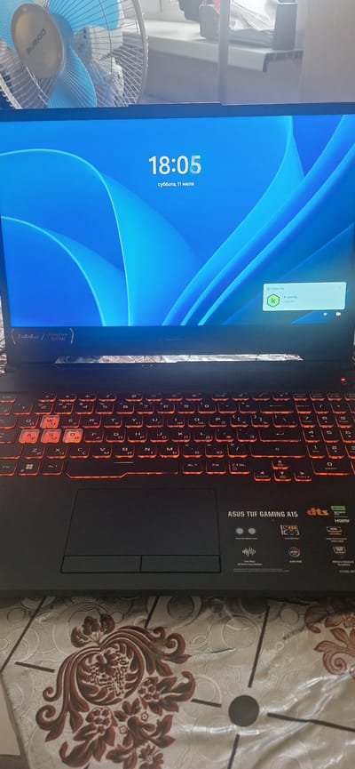 The image shows a laptop computer with a glowing red keyboard and a blue wallpaper displaying the time "18:05" and the date "суббота, 11 июля" (Saturday, July 11). A small window indicating "Kaspersky" is open in the upper right corner of the screen. The laptop appears to be placed on a patterned tablecloth. In the background, a blue fan is partially visible. The scene is not identifiable as being in Chunsky, Russia, or any other specific city.