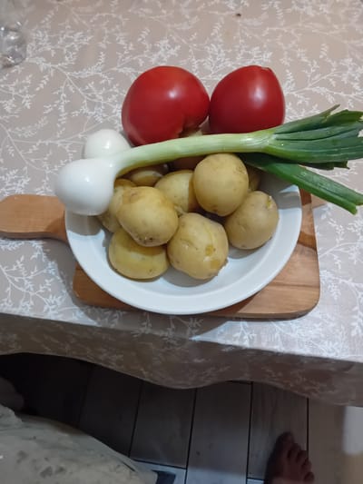 On a table covered with a light-colored tablecloth featuring a subtle white floral pattern, a white plate holds several small, yellow potatoes. Partially obscuring the potatoes is a white bulbous vegetable, likely an onion, with a long green stalk extending to the right. Behind the plate, two red tomatoes are positioned side-by-side. The plate rests on a wooden cutting board, which is itself on the tablecloth. The background shows a dark, tiled floor. There are no discernible people or actions occurring in the image, and no specific location in Dubossary, Moldova can be identified.