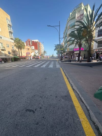 The image depicts a street scene in Lloret de Mar, Spain, characterized by a wide asphalt road with a pedestrian crosswalk. The buildings lining the street are multi-story structures with balconies, typical of a coastal tourist town. Palm trees are visible, suggesting a warm climate. On the right side of the road, a curved building with a green facade features signage that reads "St. Trop". Several people are seen walking on the sidewalk and near the shops. On the left side of the road, there are more buildings with shops and awnings. The sky is clear and blue, indicating a sunny day. The overall impression is that of a bustling commercial street in a Mediterranean resort.