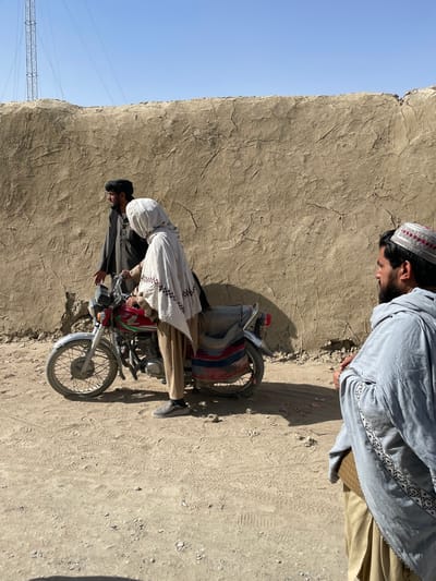 This image depicts an outdoor scene in Spin Boldak, Afghanistan, under a clear blue sky. The background is dominated by a tall, cracked mud-brick wall. The ground consists of unpaved, light brown dirt with visible tire tracks and footwear impressions. A metal lattice tower is visible in the upper left background.
Three individuals and a red Honda motorcycle are present. The motorcycle, equipped with chrome handlebars, fenders, and spokes, is centrally positioned, resting on its kickstand. A multi-colored striped fabric bundle is secured to its rear. A person, whose gender is indeterminate due to being fully covered by a light-colored patterned shawl and tan trousers, stands astride the motorcycle with both hands on its handlebars.
To the left, a man with a dark beard, wearing dark traditional attire, stands slightly behind the motorcycle. He extends his right hand with an open palm towards the person on the motorcycle, his gaze directed at them. To the right, in the foreground, another man with a dark beard wears a white and red patterned cap and a light blue patterned shawl over light brown clothing. He is seated or squatting, facing towards the central figures, with his hands interlocked in his lap. The scene captures a static moment of interaction on a dusty pathway.