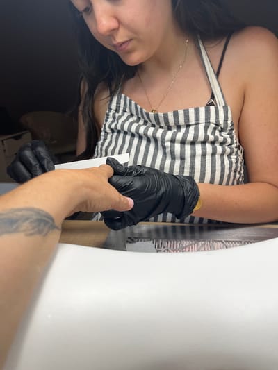 The image depicts a close-up of a person's hands engaged in a nail care activity, likely a manicure, at a salon in Montrond-les-Bains, France. The person performing the service is wearing black nitrile gloves and a striped apron over a black tank top. They are holding a white nail file and working on the fingernail of another person, whose arm, partially tattooed, is visible in the foreground. The setting appears to be a well-lit indoor space, possibly a nail salon, with a clean, white surface in the foreground that could be a sink or a table. The background is dark, suggesting controlled lighting for the task.