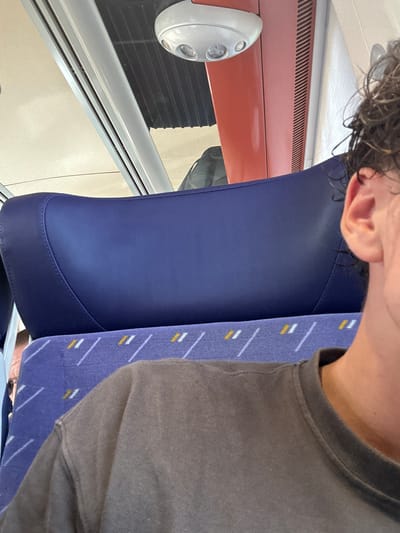 The image shows the interior of a train car, likely in the city of Agde, France, based on the context provided. The foreground is dominated by the shoulder and side of a person wearing a grey t-shirt. Behind them, a dark blue headrest of a train seat is visible. Further back, a portion of another seat with a purple fabric and white and yellow diagonal stripe pattern is seen. Above the headrest, part of a red panel is visible, containing a white light fixture with two bulbs. The overall scene suggests a person is seated on a train, looking forward or slightly to the side.