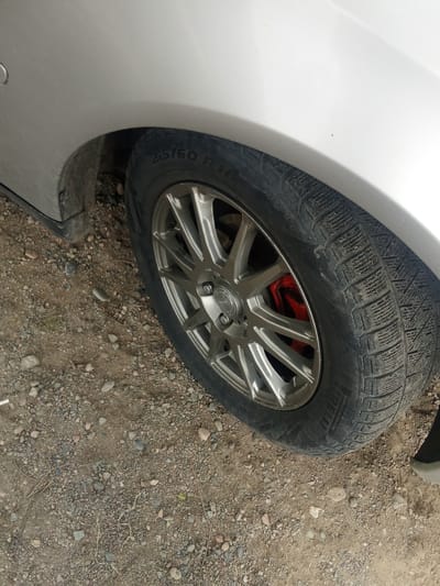 The image shows a close-up of a silver car's tire and wheel, positioned on a gravelly surface. The tire is a winter tire with prominent tread patterns and some dirt accumulation. The wheel is a multi-spoke alloy rim, and a bright red brake caliper is visible behind it. The car's body, a light silver color, is partially visible in the upper portion of the frame. The ground consists of a mix of dirt and various sizes of grey and brown gravel, with a few small twigs scattered about.

The scene appears to be outdoors, given the gravel and the natural lighting. There are no people visible in the image. The context of Alatau, Kazakhstan, suggests a potentially cold or mountainous region, which aligns with the presence of a winter tire. The lighting suggests it is daytime, but the specific time of day and weather conditions are not clearly discernible. The overall impression is a static shot of a car's wheel and surrounding environment, likely taken in a rural or unpaved area.

Visible text on the tire includes "215/60 R16" and "IRELLI", indicating the tire size and brand.