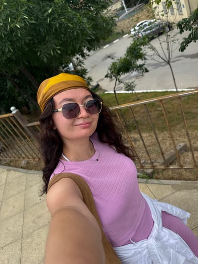 A young woman wearing a yellow and brown headscarf, large sunglasses, and a pink top is taking a selfie outdoors. She has long, dark, curly hair and is wearing a silver necklace with a small dark pendant. A white shirt is tied around her waist. In the background, there is a metal railing, trees, and a street with a parked car. The location is identified as Derbent, Russia. The overall scene suggests a casual outdoor setting.