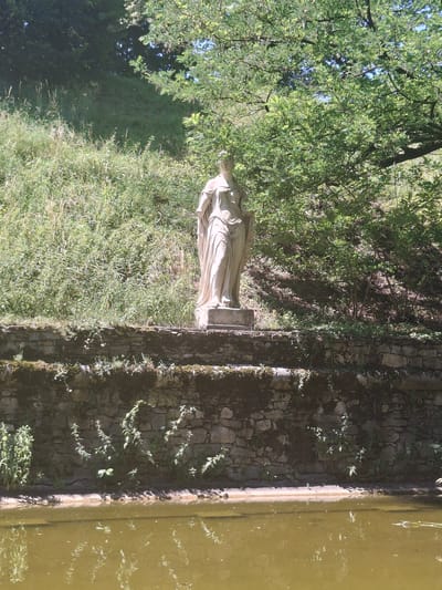 The image depicts a classical stone statue of a robed female figure standing on a stone pedestal. The statue is situated in an outdoor setting, with lush green foliage and trees in the background. In the foreground, there is a stone wall with plants growing on and around it, and a body of water, possibly a pond or canal, with reflections of the surrounding vegetation. The scene appears to be in a garden or park. Based on the provided context, the location is identified as the Volksgarten in Graz, Austria, specifically near the "Kaiser Joseph II" statue. The statue itself is likely a decorative element within the garden. The lighting suggests it is a sunny day.