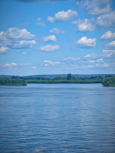 A wide, tranquil river flows through a verdant landscape under a bright blue sky dotted with fluffy white clouds. The river occupies the foreground and midground of the image, its surface rippling gently, reflecting the blue of the sky and the surrounding greenery. Along the banks, dense trees and foliage create a lush, green border that stretches into the distance. Beyond the treeline, a hazy, rolling landscape of hills can be seen under the expansive sky. The image captures a serene natural scene, likely in a rural or natural setting, such as the outskirts of Krutoy Log, Russia, known for its natural beauty and proximity to water bodies.
