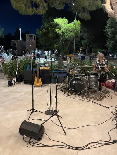 The image depicts an outdoor nighttime scene, likely at a public gathering in Toritto, Italy. In the foreground, a drum set with the "ACUSTICA" logo is set up on a rug, and a person with gray hair is playing the drums, wearing a black and white patterned shirt. To the left of the drum set, a blonde electric bass guitar stands on a stand next to a black amplifier labeled "RCF." Several microphone stands are positioned in front of the drum set and towards the left side of the frame. A black, rectangular stage monitor speaker is placed on the ground in the lower left corner. In the background, several large speakers are mounted on stands. Behind the stage equipment, a crowd of people are visible, some seated and others standing, with trees and vegetation forming a natural backdrop. A tall, white monument or sculpture can be seen in the distance against a dark blue, twilight sky. The ground appears to be a paved surface, possibly a plaza or courtyard.