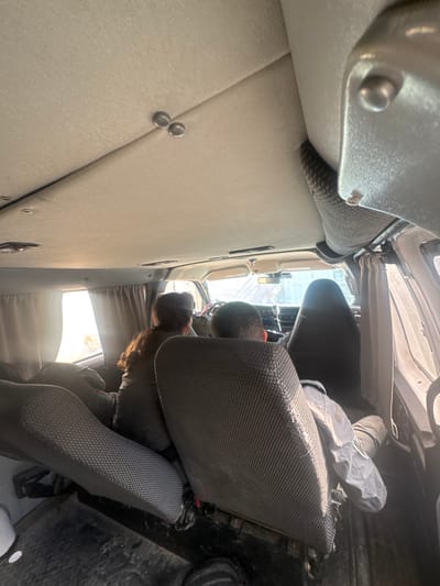 The image displays the interior of a vehicle, likely a van or large SUV, captured from a rear passenger perspective looking forward, in the city of Lod, Israel. The light-colored, fabric-lined ceiling features two metallic attachment points and overhead lights. Three individuals are visible, all facing forward. In the front passenger seat, a person with long, dark-brown hair is seated. The driver is partially visible behind the steering wheel to the left of the front passenger. In the middle row, a third individual, presumed male, is seated with their back to the camera, wearing a dark jacket. The seats are upholstered in dark gray, textured fabric. Side windows are fitted with light-colored curtains, partially open. Bright daylight illuminates the interior from the windows and front windshield. The front windshield shows a blurred view of an exterior environment, possibly an urban or industrial structure with vertical lines. A rearview mirror is visible at the top center of the windshield, and a dark seatbelt mechanism is present on the upper right interior. The floor is dark and utilitarian.