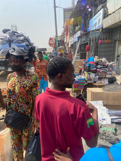 This image captures a bustling outdoor market street in Igbede, Nigeria, under bright daylight conditions.
In the foreground, a male individual is observed from behind, wearing a red short-sleeved polo shirt with a green "RMN" logo on the right sleeve. He holds a dark-colored smartphone in his right hand. To his left, a female individual walks, facing partially away from the camera, wearing a multi-colored leopard-print patterned jumpsuit and a black face mask. She carries a large, irregularly shaped bundle of items, wrapped in clear plastic and dark material, balanced on her head. A black shoulder bag is worn across her body. A hand from another individual is visible touching the back of the male in the red shirt.
In the midground, a male individual wearing an orange and black patterned shirt and blue trousers walks towards the right side of the frame. A white vehicle, possibly a truck or van, is partially visible on the left, displaying a Nigerian license plate with the partial identifier "71JGS". Several other individuals are present in the midground, moving through the scene.
The right side of the frame features numerous market stalls and commercial buildings. Stacks of folded clothing items in various colors are displayed on tables or makeshift stands. Boxes, electronic components, and a metal fan guard are also visible for sale. The buildings in the background are multi-storied with exposed metal structures, featuring advertising signs such as a yellow billboard reading "Settle your nwa boi," and another sign stating "FIRST CHOICE LGP." A "SOLAR" sign is also visible. Clothes, including a red jersey with the number "23," hang from racks. Streetlights and overhead wiring are present.
The ground surface appears unpaved and dusty. The sky is clear, suggesting a sunny day, with some atmospheric haze visible in the distance. The overall environment indicates a busy commercial street with active pedestrian traffic.