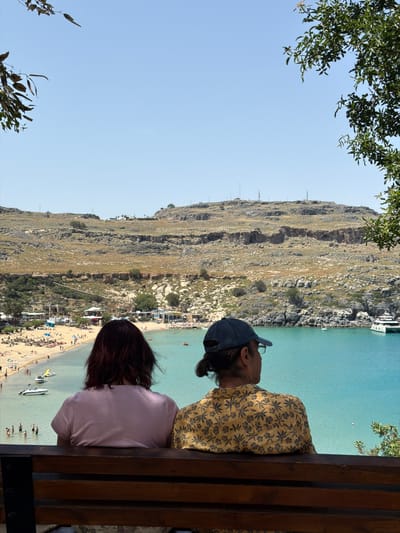 A medium shot shows two people sitting on a wooden bench looking out over a beach in Lindos, Greece. The person on the right, wearing a yellow shirt with a dark floral print and a blue baseball cap, appears to be male. The person on the left, wearing a light pink shirt, has dark brown hair and appears to be female. In the background, there is a crowded beach, turquoise water, and a large, rocky hill. A few green leafy branches are visible in the upper left and right corners of the image. The sky is a clear, light blue.