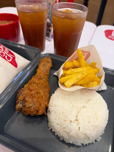 This media file captures a close-up view of a meal presented on a dark grey tray. The meal consists of a generous portion of fluffy white rice, a serving of golden-brown fried chicken, and a small white paper cone filled with french fries, dusted with a yellow seasoning and herbs. Two tall, clear glasses filled with a light brown, possibly iced tea, beverage are visible on the left side of the frame, suggesting a refreshing drink accompanies the meal. A hint of a red cup can be seen at the bottom left. The background is blurred but appears to show stacked white paper items, possibly more containers or packaging, with faint red branding visible on some. The lighting suggests an indoor setting, possibly a fast-food restaurant or a casual dining establishment. The overall presentation is neat and appetizing, indicating a typical meal being served. Visible text on the paper cone and possibly on the background items appear to be part of a brand logo, with the word "CK" clearly legible in a stylized font on a white paper lining at the bottom. The location context of Dagupan, Philippines, implies this is a common meal option available in that region. There are no people or animals visible in the frame, and the focus is solely on the food and drinks. The image appears to be taken from a slightly elevated angle, looking down at the tray.