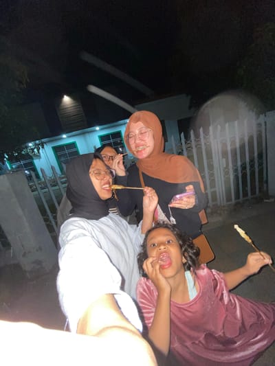 In Tanggul, Indonesia, at night, a group of four people is captured in a selfie. Two adult women, one wearing a black hijab and a striped shirt, and another wearing a brown hijab and a black shirt, are on the left and right respectively. Both are smiling and holding skewers of food, with the woman in the brown hijab also holding a pink phone. In the foreground, a young girl in a pink velvet dress is also holding a skewer of food and looking at the camera with her mouth open. Another person, a woman wearing glasses and a black hijab, is partially visible behind the woman in the black shirt. The background shows a building with green-trimmed windows and a white fence. The scene appears to be a casual outdoor gathering.