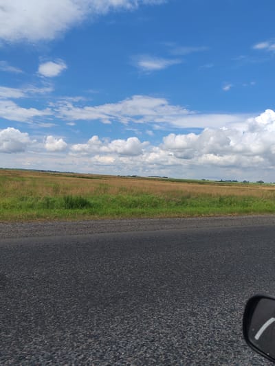 The image depicts a rural landscape under a bright blue sky with fluffy white clouds. In the foreground, a dark asphalt road stretches across the bottom of the frame. Beyond the road lies a vast field of dry grass and some green vegetation, with a gentle slope upwards towards a distant horizon. The landscape appears flat and open, characteristic of the Урыцкі сельскі Савет region in Belarus. The scene suggests a clear, possibly summer day.