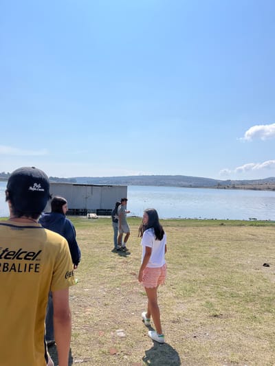 The image depicts an outdoor scene in El Oasis Valsequillo, Mexico. A body of water borders a grassy area where several people are present. A young woman wearing a white shirt and pink skirt stands in the foreground. Behind her, a man in a grey shirt and shorts stands near a silver building. In the foreground, a person wearing a black baseball cap and a yellow shirt are seen from the back. Another person wearing a blue jacket is also partially visible. The sky is blue with a few clouds.