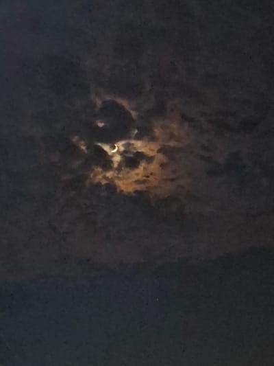 An image captures a partial solar eclipse as seen through clouds. The moon obscures a portion of the sun, creating a crescent shape of light. The clouds are dark and textured, with some areas illuminated by the sun's rays peeking through. The bottom portion of the image is a dark, blurry expanse of sky, indicating a low-light environment, likely dusk or pre-dawn. The scene appears to be outdoors, possibly in Yovon, Tajikistan, given the context provided. No people or other specific objects are visible in the frame.