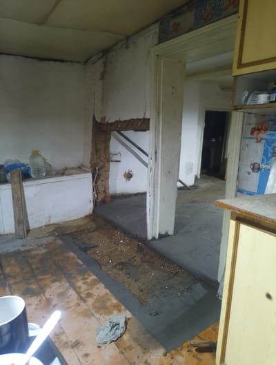The image shows an interior scene of what appears to be a room undergoing renovation or repair. The setting is rustic and somewhat dilapidated. The floor in the foreground is made of wooden planks, with patches of fresh gray cement poured in a section, suggesting ongoing construction work. Some debris and dirt are scattered on the floor.
To the left, there is a white counter or shelf unit against a wall, with several large plastic bottles of liquid on top. A wooden plank stands upright against the counter. The walls are rough and unfinished in places, particularly around an opening that leads to another room. Exposed beams or structural elements are visible in the wall of this opening.
The doorway in the center leads into a dimly lit hallway or another room, with a glimpse of furniture in the distance. On the right side of the image, part of a wooden cabinet or shelf is visible, with yellow-painted doors and a wooden trim. Above this cabinet, a section of wallpaper with a floral pattern is visible.
The overall lighting suggests an indoor environment, likely during the day but without strong natural light. The scene conveys a sense of unfinished work and a potentially older building. There are no people visible in the image. The location context provided is "Бородулиха ауылдық округі, Kazakhstan," which suggests this is a rural area in Kazakhstan. No visible text is present in the image.