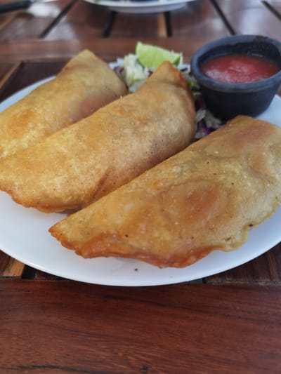 Three golden-brown empanadas are arranged on a white plate, with a small bowl of red salsa beside them. The empanadas are fried and have a crispy texture. There is also a small amount of shredded lettuce and a lime wedge present. The scene is set on a wooden table. This location, with the provided coordinates, is a street food vendor in Unidad de Riego, Mexico.