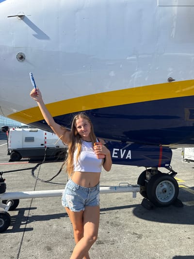 A young woman stands on an airport tarmac, posing in front of a large airplane. She is wearing a white crop top and denim shorts, and is holding an orange smartphone in her right hand and a blue pen in her left hand, which is raised above her head. The airplane is predominantly white with a yellow and dark blue stripe along its side, and the letters "EVA" are visible on its fuselage. In the background, a white service vehicle with the "GUINAULT" logo is parked. The ground is paved with asphalt, and shadows are cast by the airplane and other objects. The image appears to have been taken on a sunny day. The location of Kruja, Albania is not depicted in this image.