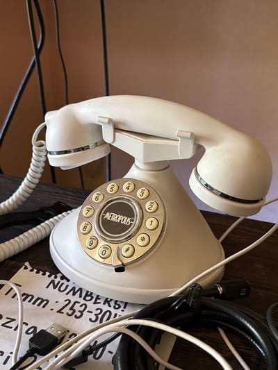 A white rotary dial telephone with a cream-colored handset is centrally positioned on a dark wooden surface. The telephone features a chrome dial with numbers and letters, and a circular metal plate in the center displaying the word "METROPOLIS". A coiled white cord connects the handset to the base. To the left of the telephone, a black strap is visible. In the foreground and to the right, various cables, including white and black wires, are scattered across the surface. A torn piece of paper with handwritten text that reads "NUMBERS! mm: 253-30" and "Ezro" is partially visible in the lower left corner, along with a USB connector. The background is a plain, light brown wall, with vertical dark lines, possibly representing a rack or stand, behind the telephone. The overall scene is a close-up shot of the telephone and surrounding cables and paper.