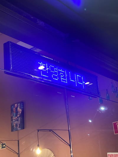 A vertically oriented blue LED sign displays the text "무료주차" (Korean for "Free Parking") against a dark background, likely at night. The sign is mounted on a wall or structure, potentially indicating a parking area or business in Caxias do Sul, Brazil. The scene also features a blurred backdrop with a brown or beige wall, light fixtures, and other miscellaneous items, including a picture on the wall.