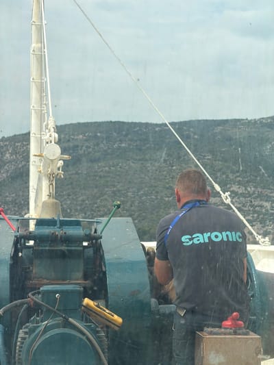 A medium shot shows a man working on machinery, likely on a ship, against a blurred background. The man is seen from behind, wearing a dark t-shirt with "saronic" printed on the back, indicating possible employment with Saronic Ferries or a similar organization operating in the Saronic Gulf. He has short, gray hair, and part of a blue lanyard or ID card is visible around his neck.

The machinery is dark teal with corroded, worn details, suggesting exposure to marine elements. An electric motor is partially visible. Levers and wires, including a handheld yellow device with black switches, are attached. A red valve handle is visible near the man's waist.

Outside the machinery is a blurred coastal landscape with green vegetation and hilly terrain. The location appears to be the Municipal Unit of Methana, Greece, based on the man's shirt indicating maritime employment in the Saronic Gulf region. The image captures an environment where someone is actively engaged in ship maintenance or repair.