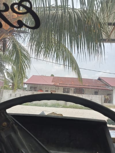The image depicts a view from inside a vehicle, looking out through a windshield adorned with decorative lettering and palm fronds. In the foreground, the top of a steering wheel is visible. Beyond the windshield, a building with a red corrugated metal roof is the primary subject. The building appears to be a single-story structure with multiple windows and a wall in front of it. The surrounding environment consists of a sandy or dirt road, some greenery, and a cloudy sky. The image is likely taken in a tropical or subtropical region, as suggested by the palm trees. While the exact location of Melayu Besar Kota, Indonesia is not explicitly shown, the presence of tropical vegetation and architecture aligns with the general landscape of such areas. There are no individuals or direct human actions visible in the frame.