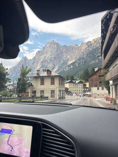 The image captures a view from inside a vehicle, looking forward through the windshield. A modern car's dashboard and navigation screen are visible in the foreground. The exterior scene shows a street in the town of San Vito di Cadore, Italy, with traditional alpine architecture. Mountains, covered in trees, dominate the background under a cloudy sky. Several buildings line the street, including a light-colored building with red shutters and a prominent red-tiled roof in the mid-ground. A road sign indicating "92 SS 51" is present. Cars are parked along the street, and some outdoor seating areas are visible. The street appears wet, suggesting recent rain. There are no people clearly visible in the image.
