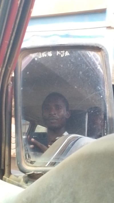 The image depicts a close-up view of a vehicle's driver's side mirror, reflecting a dark-skinned male occupant. The scene is located in Igbo Ukwu, Nigeria. The mirror surface, appearing aged and somewhat obscured by dust or scratches, displays the alphanumeric identifier "355 KJA" across its upper portion. Reflected within the mirror is an adult male with short dark hair and a light beard, wearing a light-colored collared shirt. His right hand is visible within the reflection, holding a dark rectangular object, consistent with a mobile phone. The immediate foreground, out of focus, reveals light-colored interior components of the vehicle, possibly a dashboard or door panel, with a red vertical element visible on the far left, indicating the vehicle's structure. The reflected background beyond the individual is indistinct, suggesting an outdoor environment, with a hazy light-blue horizontal element visible above the mirror frame.