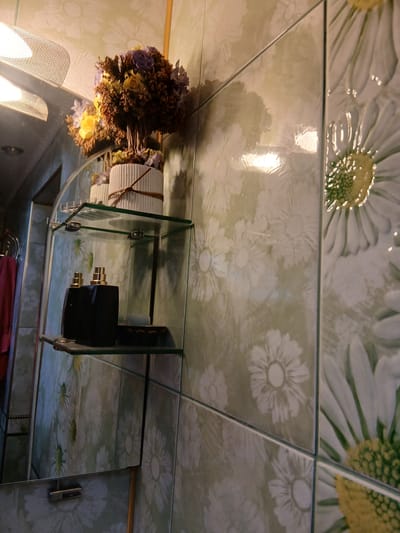 A close-up, eye-level shot shows a glass shelf with two black perfume bottles and a small box. Above the shelf, a bouquet of artificial flowers in a white ribbed pot is visible. The background features light green tiles with a floral pattern of daisies. A reflective surface, likely a mirror or polished wall, is to the left, showing a blurred reflection of a light fixture and a red towel. The overall scene appears to be in a bathroom.