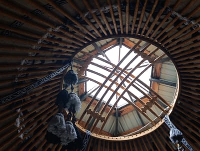 This image shows the interior of a yurt in Kegeti, Kyrgyzstan. The perspective is looking upwards towards the circular opening at the top of the yurt, known as the 'tunduk'. The frame of the yurt is constructed from numerous wooden poles radiating outwards and upwards, creating a radial pattern. These poles are bound by rope and fabric elements, with decorative tassels hanging from some points. The interior walls are covered with textiles, featuring geometric patterns in black and white. The 'tunduk' is open to the sky, allowing natural light to enter, and reveals a secondary wooden frame structure supporting the opening. The overall impression is of a traditional nomadic dwelling, highlighting the structural elements and decorative details of the yurt.