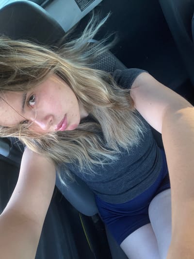 This image presents a close-up selfie of a single female individual situated inside a vehicle. The specific location within Santa Rosa, Argentina, cannot be determined from the visual information provided.
The subject is a young adult female with light skin, long wavy blonde hair that appears darker at the roots, and brown eyes. Her head is tilted slightly to her left, and her gaze is directed towards the camera. She is dressed in a dark gray, ribbed, short-sleeved top and dark blue shorts. Minor blemishes are discernible on her lower left facial area.
The background consists of the dark interior of a vehicle, including a dark gray car seat with a textured fabric pattern. A white label containing text is partially visible on the upper right internal panel of the vehicle. Natural light, consistent with sunlight, illuminates the subject from the viewer's right, creating bright highlights on her hair and left arm. The individual's right arm is positioned to operate the camera, while her left arm is visible resting on her lap. The upper thighs are visible above the dark blue shorts.