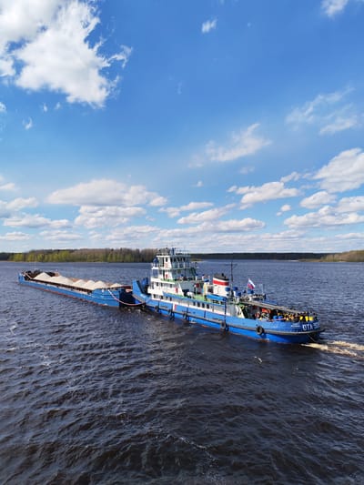 A blue and white tugboat, labeled "OTA-929" and "Санкт-Петербург" (Saint Petersburg) on its hull, is pulling a barge loaded with a light-colored granular material across a body of dark water. The tugboat is moving, as evidenced by the wake behind it. A Russian flag is visible on the tugboat. On the barge, the granular material is heaped into several mounds. The shoreline is visible in the background, with a line of trees. The sky is blue with scattered white clouds. There are no people distinctly visible. The location is not specified as Pochinok, Russia in the image.