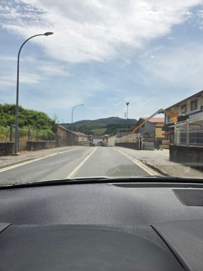 The image depicts a view from inside a vehicle, looking out through the windshield at a street scene in Doneztebe / Santesteban, Spain. The street is paved and appears to be a two-lane road with white lane markings. On the left side of the road, there is a sidewalk, a stone wall, and a lush green hillside covered in trees. A tall street lamp with a curved pole stands on the left side of the road. On the right side of the road, there are buildings. One building features an extended yellow crane with a basket at the end, suggesting some form of construction or maintenance work is in progress. The sky above is partly cloudy with a clear blue sky visible. In the foreground, the dashboard of the vehicle is visible, with a textured surface. The overall impression is a quiet, somewhat rural street in a Spanish town on a bright day.