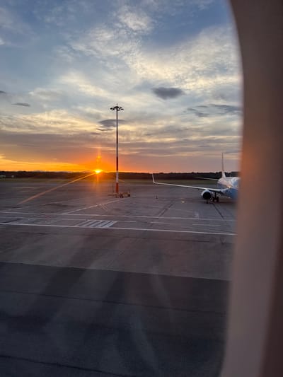 An airplane is parked on the tarmac of an airport in Somma Lombardo, Italy, as the sun sets. The sky is filled with dramatic clouds, tinged with the orange and yellow hues of the setting sun. The sun is a bright orb, low on the horizon, casting long shadows across the asphalt. The airplane, a light blue and white passenger jet, is positioned on the right side of the frame. A tall light pole with red and white markings stands in the middle of the tarmac, closer to the foreground. The foreground is dominated by the dark asphalt of the airport, marked with white lines indicating taxiways or parking spots. The background shows a line of trees and distant hills, silhouetted against the vibrant sky. The image is taken from the perspective of someone inside an aircraft, looking out a window, as evidenced by the blurred, light-colored frame on the right side of the image.