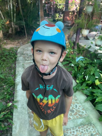 A young boy wearing a blue helmet with cartoon bear ears and a brown t-shirt with an AC/DC lightning bolt graphic sticks his tongue out at the camera. He is standing on a walkway made of concrete and embedded stones, flanked by lush green foliage and bamboo plants. The walkway appears to be in an outdoor setting, possibly a garden or park, in Hoi An, Vietnam, suggested by the tropical vegetation and the architectural style of a blurred building in the background. The lighting suggests it is daytime, with dappled sunlight filtering through the leaves.