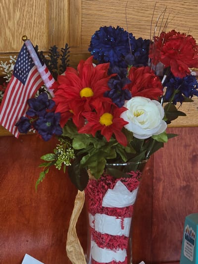 A close-up, eye-level shot shows a patriotic floral arrangement, likely for a holiday such as the Fourth of July or Memorial Day, placed against a backdrop of wooden paneling. The arrangement is composed of red, white, and blue artificial flowers, including large red daisies with yellow centers, deep blue clusters, and a white rose. A miniature American flag on a stick stands prominently to the left of the flowers, with its upper tip adorned with a small gold ball.
The flowers are contained within a clear glass vase decorated with red and white crinkle-cut paper filler, giving it a festive appearance. A tan ribbon is tied around the base of the vase, and white tissue paper is layered over the red filler in bands. The vase sits on a surface that is partially visible, and to the right, a teal-colored box with the word "BALLOON" and images of balloons is visible, suggesting a party or celebration. The lighting appears to be natural, casting soft shadows. The overall impression is one of festive decoration. The specific location in Cumming, United States of America, is not identifiable from the image.